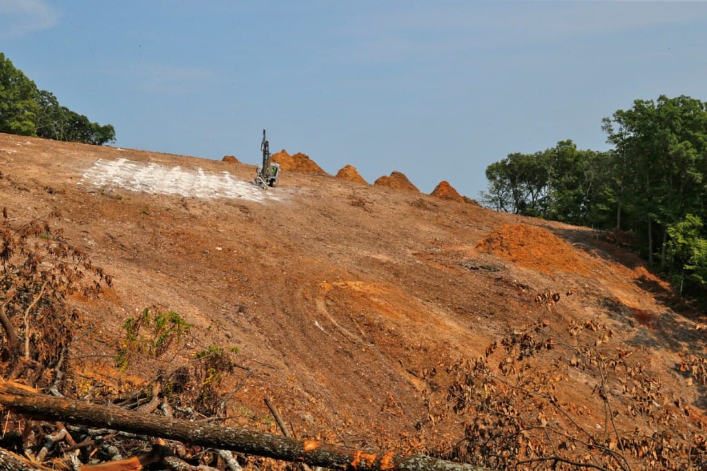 A drilling rig from R&K Excavators prepares holes to accept the explosive charges for blasting to move soil and rock down the hillside. (08/22/18)
